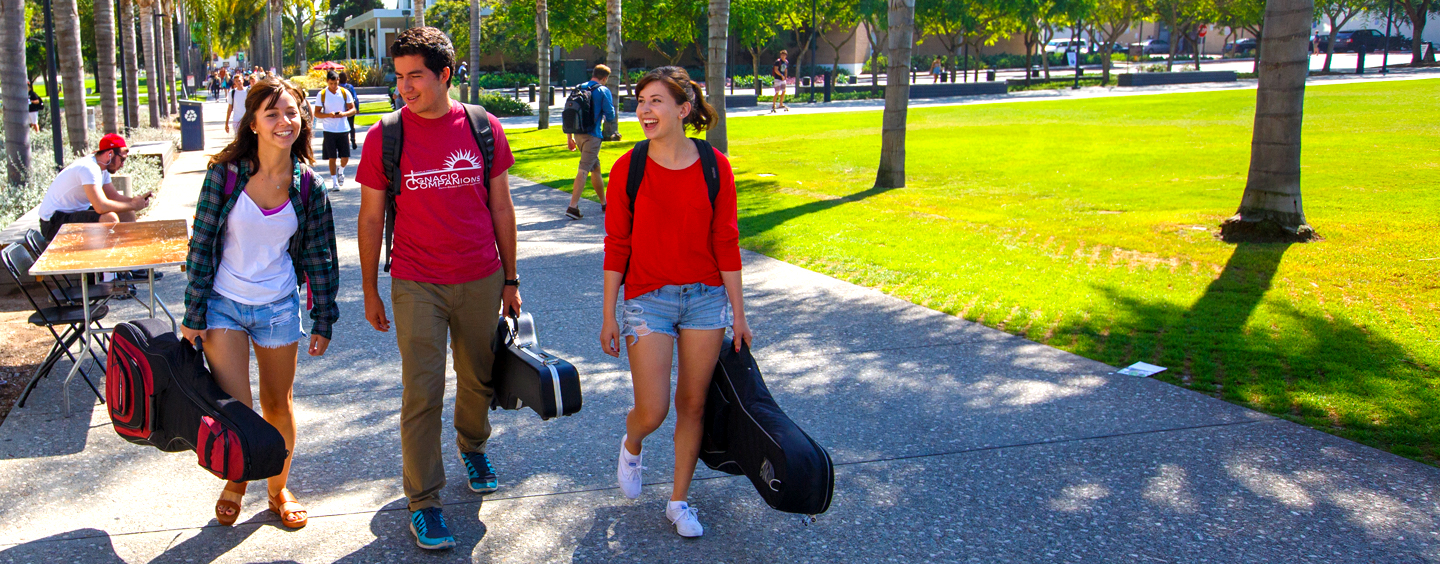 Three students carrying guitars walk around campus.