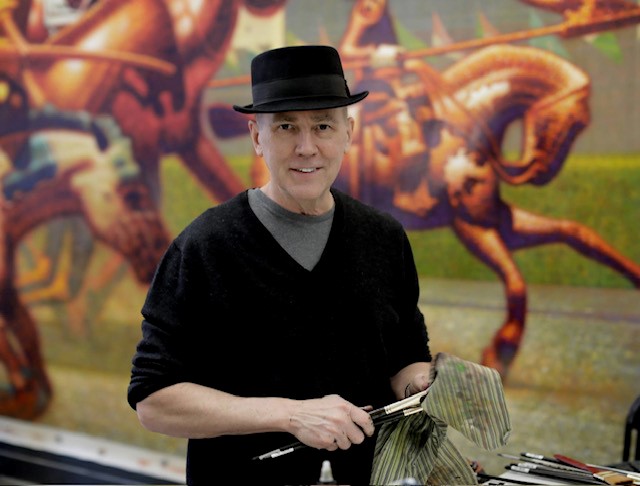 Photo of a man wearing a black sweater and hat while standing in front of a painting of running horses.