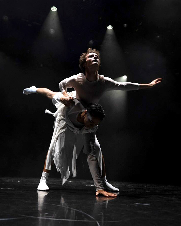 Two dancers in white under the spotlights of a darkened stage.
