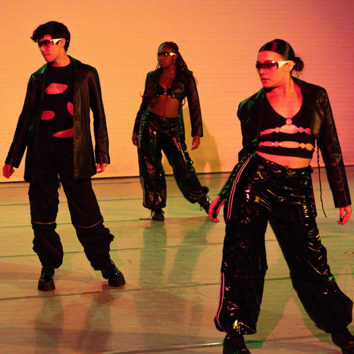 Three dancers in dark outfits in front of a yellow-orange background.