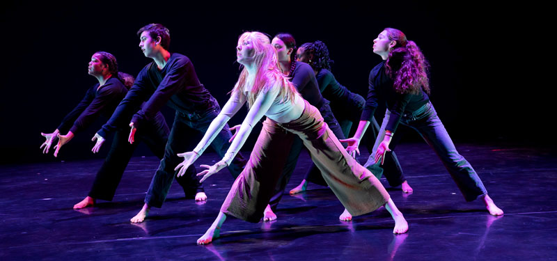 A group of dancers performing onstage under a magenta light.