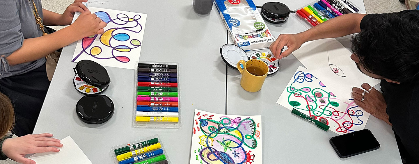 Art supplies scattered across a table as three people work on art projects