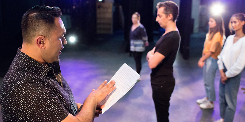 Director reading a script on stage with actors in the background