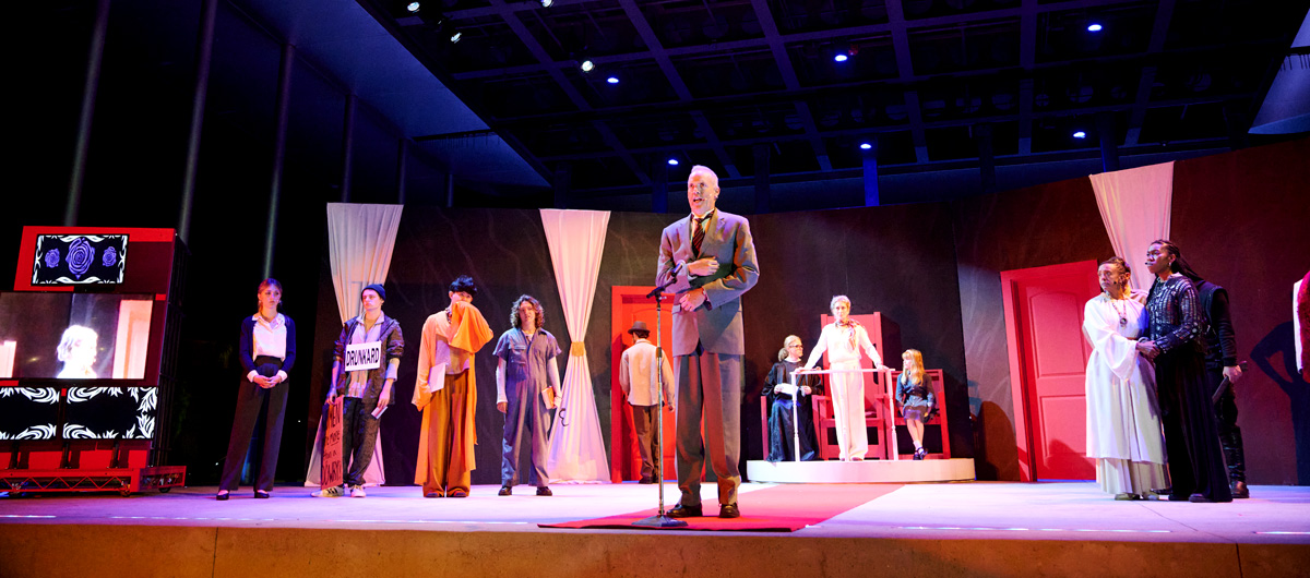 A man in a suit speaking to the audience while others look on.