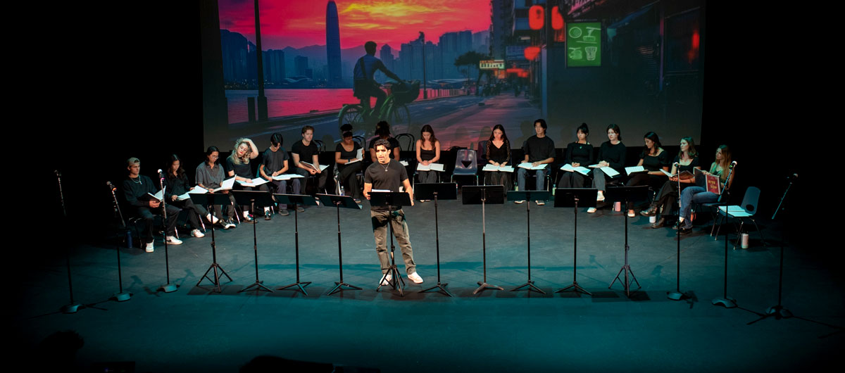 A man stands alone on stage, while the others remain seated behind them.