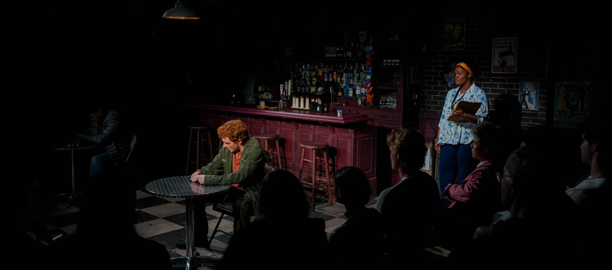 A man sits alone at a bar while a woman looks on from a distance.